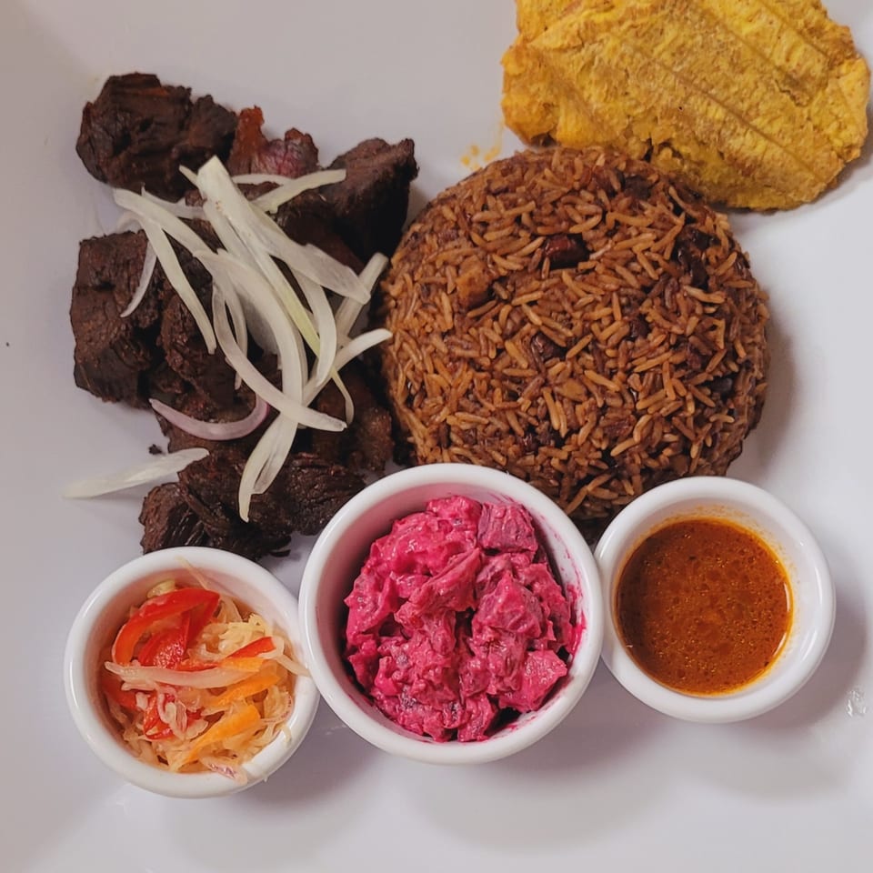 Haitian griot with rice and beans, fried plantain, pikliz, beet salad, and sauce on a white plate.