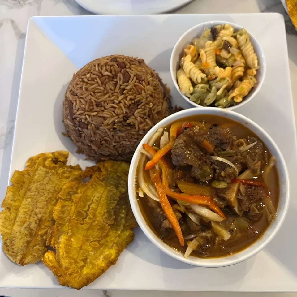 Haitian meal with rice, beef stew, pasta salad, and fried plantains on a white plate.