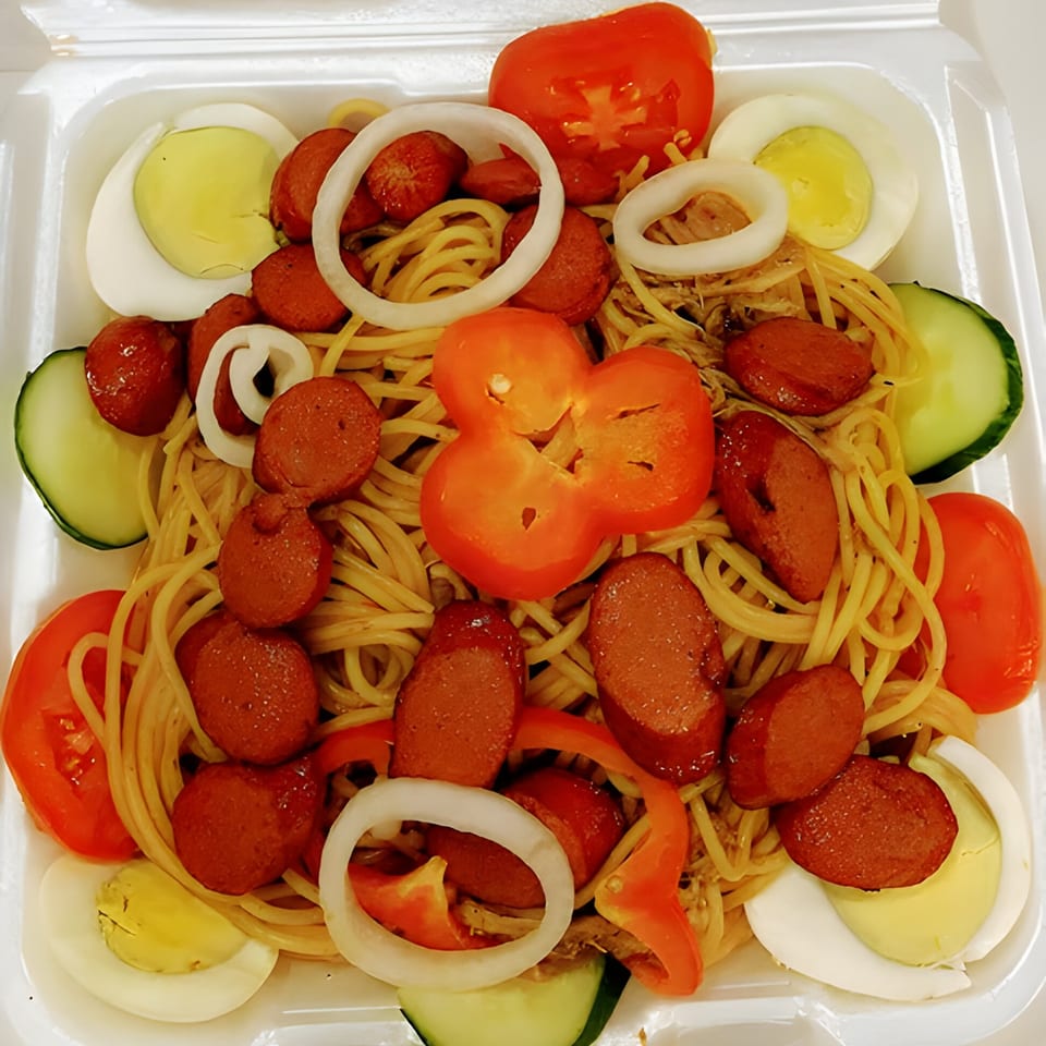 Haitian spaghetti with sliced hot dogs, hard-boiled eggs, tomatoes, cucumbers, and onions in a white container.